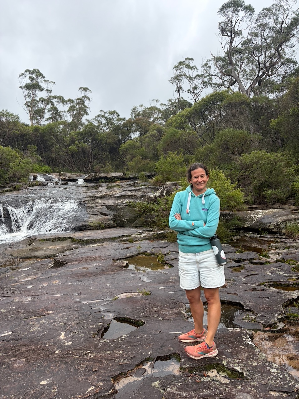 Steph standing on wet sandstone rocks at Carrington Falls with water flowing behind her.