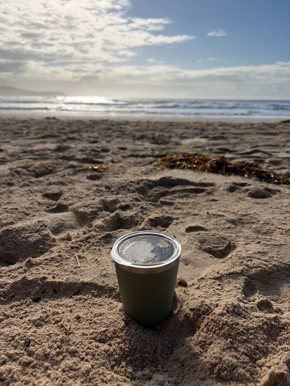 A coffee cup sitting in the sand on the beach at Killalea with the ocean in the background.