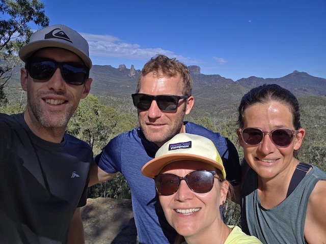 Four people smiling in a selfie at a rocky lookout with the Warrumbungle formations silhouetted behind them.