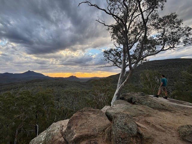 A person standing on a rocky outcrop at dusk with the Warrumbungle volcanic formations silhouetted on the horizon and the sky turning orange.