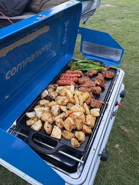 A camp griddle loaded with sausages, steak, roast potatoes and vegetables cooking outside.