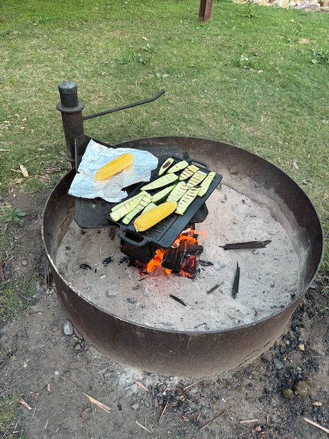 Corn cobs and zucchini cooking over an open campfire in a cast iron pan, flames glowing below.