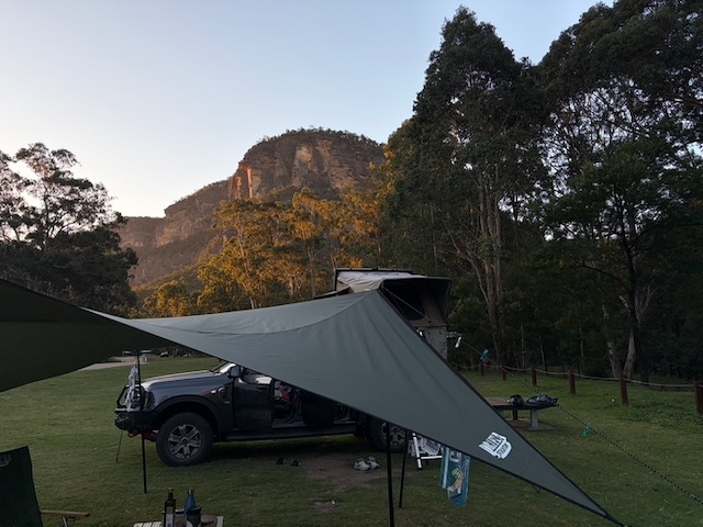 Roger the Ranger under the tarp awning at Camp Blackman, with a tall volcanic rock spire rising sharply above the treeline.