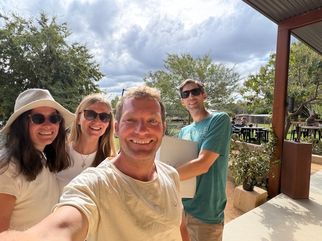 Group selfie of four people smiling outside a winery building with gum trees and outdoor seating behind.