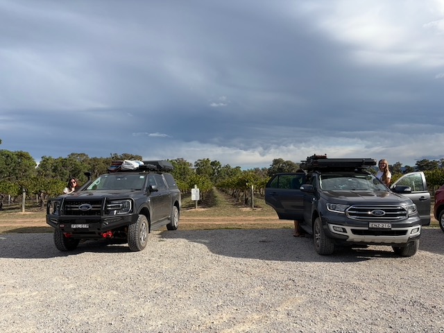 Two 4WD vehicles with rooftop tents parked in a winery car park with vineyards and dramatic clouds behind.