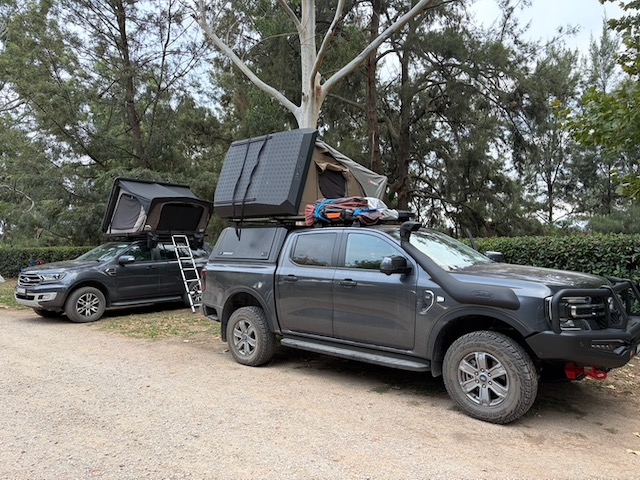 Two 4WD vehicles with rooftop tents parked side by side at Coorongooba campground, with tall trees behind.