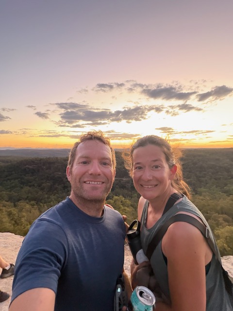 Matt and Steph selfie at the Pilliga sunset viewpoint, with glowing orange and pink sky behind them and endless forest below.
