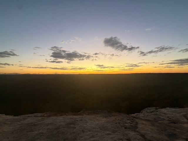 Wide view from the Pilliga Pottery lookout rock ledge at sunset, with a pale orange sky fading to dark over a vast flat forested plain.