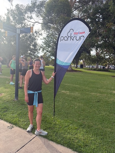 Steph smiling and holding the Mudgee parkrun banner on a sunny morning.