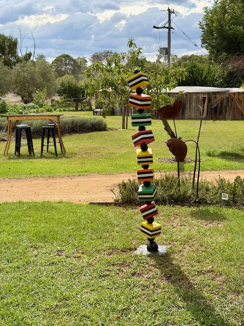 A colourful stacked geometric sculpture in the Rosby winery garden under a cloudy sky, with vines and buildings behind.