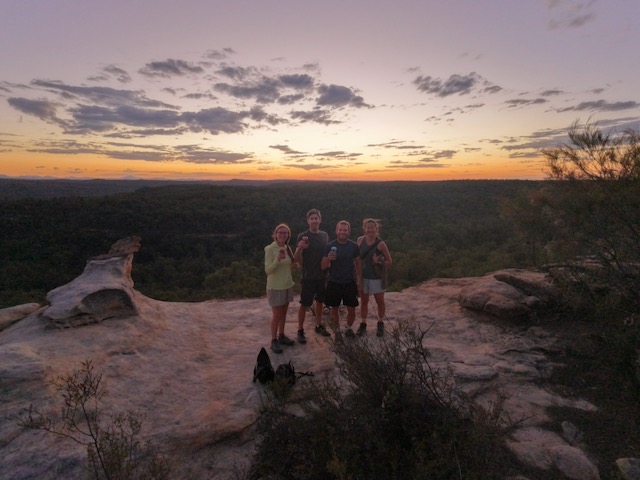 Four people standing on a sandstone ledge at the Pilliga Pottery viewpoint, drinks in hand, with a vast forested landscape stretching to the horizon under a deep purple and orange sunset sky.